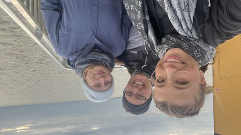 drei junge Frauen in warmer Kleidung stehen auf einem Boot und lächeln in die Kamera. Im Hintergrund ist das Meer zu sehen.