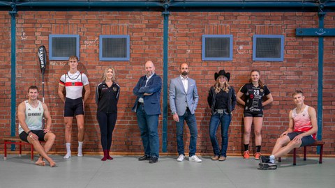 Gruppenbild mit sechs Sportstipendiat:innen, Professor Odenbach und Marko Schimke (DHSZ) in einer Turnhalle vor einer Backsteinmauer