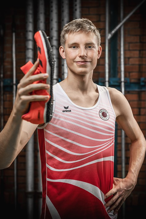 Porträtaufnahme. Tom Liebscher im Sportdress mit Padel in der Hand. Lächelt in die Kamera. Im Hintergrund Backsteinwand mit senkrechten Rohren