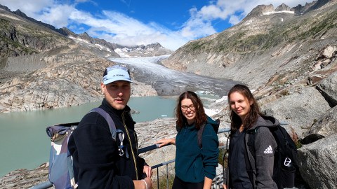Auf dem Foto ist im Hintergrund eine karge, alpine Berglandschaft zu sehen. In der Mitte mündet eine Gletscherzunge in einen See. Davor posieren drei Personen in Wanderkleidung, links ein junger Mann, neben ihm zwei junge Frauen.