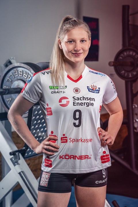Porträt Sina Stöckmann. Die Sportlerin hat ein weißes T-Shirt mit Sponsoren-Logos sowie eine kurze schwarze Short an. Sie hat die Hände in die Hüften gestemmt und lächelt in die Kamera. Ihre blonden Haare sind zu einem Pferdeschwanz gebunden. Hinter ihr ist ein Fitness-Raum zu erkennen.