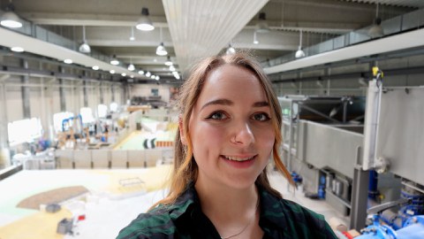 Auf dem Foto ist eine lächelnde junge Frau mit zusammengebundenen hellbraunen Haaren zu sehen. Im Hintergrund sieht man eine große Labor-Halle