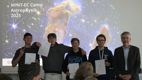 Vier Jugendliche und Prof. kobel stehen nebeneinander vor einem Hintergrund der "die Säulen der Schöpfung" zeigt mit der Aufschrift MINT-EC CAmp Astrophysik 2025". Die Jugendlichen halten Teilnahmeurkunden.