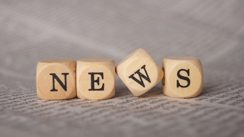 Photo with four wooden cubes lying on a printed sheet of paper. On each cube is a letter. The letters make the word "News".
