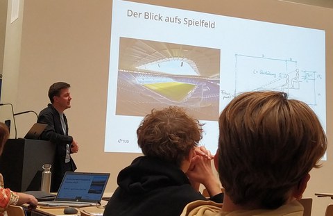 Zwei Schüler sitzen im Vordergrund, vor einer Präsentation über ein Stadiondach