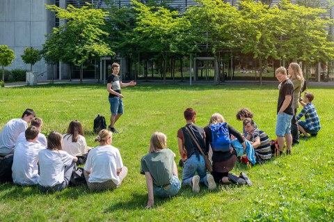Foto. Eine Gruppe von Personen sitzt auf einer Wiese. Im Hintergrund ist ein Gebäude zu sehen, vor dem Bäume stehen. 