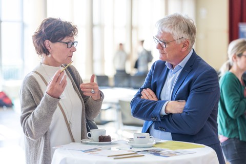 Ein Mann und eine Frau unterhalten sich an einem Stehtisch. AUf dem Tisch liegen Kaffe, Kuchen und Flyermaterial.