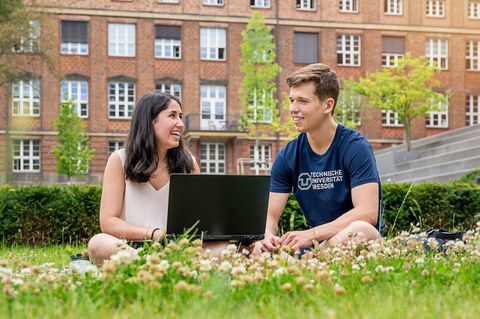 Bild von zwei Studenten, die lachend auf der Wiese zusammen am Laptop arbeiten.