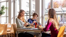 Das Foto zeigt drei Studierende beim Mittagessen. Sie sitzen an einem Tisch in der Mensa. Man erkennt einen Teller mit Nudeln. Außerdem steht ein Getränk auf dem Tisch.