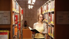 Das Foto zeigt eine Studentin in der Bibliothek. Sie steht zwischen den Bücherregalen, hält ein aufgeschlagenes Buch in der Hand und lächelt in die Kamera.