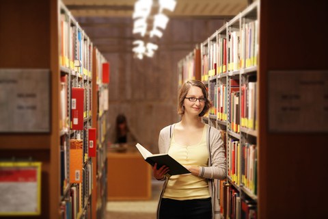 Das Foto zeigt eine Studentin in der Bibliothek. Sie steht zwischen den Bücherregalen, hält ein aufgeschlagenes Buch in der Hand und lächelt in die Kamera.