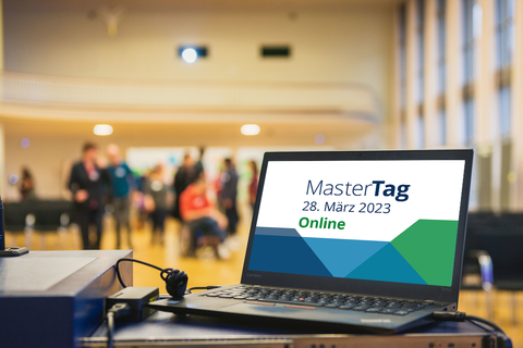 Das Foto zeigt einen Veranstaltungssaal. Im Vordergrund erkennt man einen Laptop auf dem Rednerpult. Im Laptop befindet sich ein Bild mit farbigen Kacheln und dem Schriftzug MasterTag 28. März 2023 Online