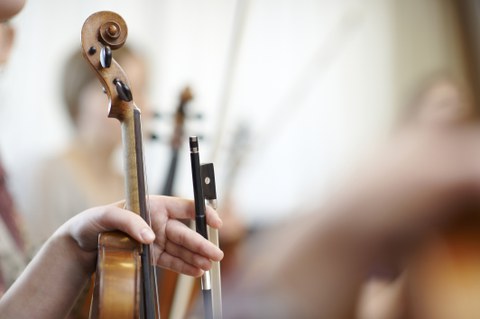 Auf dem Foto sieht man einen Ausschnitt aus einem Orchester. Im Fokus ist eine Geige, die von einer Person in der Hand gehalten wird. 