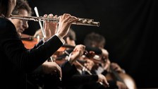 Auf dem Foto sieht man einen Ausschnitt aus einem Orchester. Im Fokus ist dabei eine weibliche Flötistin, die eine Querflöte hält. 