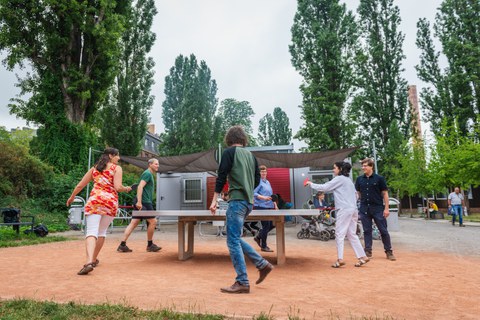 Foto: Eine Gruppe von sechs fröhlichen Studierenden spielt Tischtennis vor dem Grill Cube am Hörsaalzentrum. im Hintergrund sind Bäume zu erkennen.