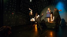 Foto: Eine Gruppe von Schauspielern spielt Theater in einem abgedunkelten Raum. Im Zentrum des Bildes steht eine Sängerin auf einer Bühne. 