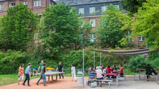 Foto: Eine Gruppe von sechs fröhlichen Studierenden spielt Tischtennis vor dem Grill Cube am Hörsaalzentrum. im Hintergrund sind Bäume zu erkennen.