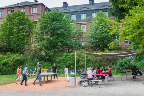 Foto: Eine Gruppe von sechs fröhlichen Studierenden spielt Tischtennis vor dem Grill Cube am Hörsaalzentrum. im Hintergrund sind Bäume zu erkennen.