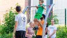 Ein Foto eines Jungen im Freien an einer Reckstange. Ein Mann hält ihn an den Beinen. Im Hintergrund stehen weitere Personen, die die Szene beobachten. Im Hintergrund Zaun, Gebäude und Grün.