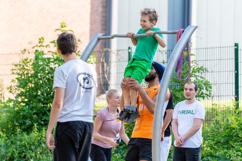 Ein Foto eines Jungen im Freien an einer Reckstange. Ein Mann hält ihn an den Beinen. Im Hintergrund stehen weitere Personen, die die Szene beobachten. Im Hintergrund Zaun, Gebäude und Grün.