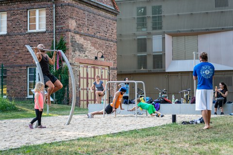 Foto: Mehrere Studenten machen Sport auf einem kleinen Sportplatz. Im Hintergrund Gebäude und Wiese. 