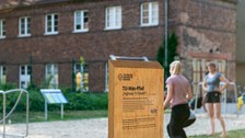 Foto eines freistehenden Holzschildes. Auf dem Schild steht "TU-Was-Pfad" und darunter "Highway TU Health" sowie weiterer nicht erkennbarer Text. Im Hintergrund ein Gebäude, Sportgeräte und zwei Personen in Sportkleidung.