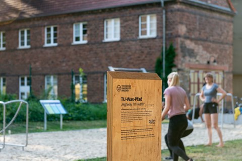 Foto eines freistehenden Holzschildes. Auf dem Schild steht "TU-Was-Pfad" und darunter "Highway TU Health" sowie weiterer nicht erkennbarer Text. Im Hintergrund ein Gebäude, Sportgeräte und zwei Personen in Sportkleidung.