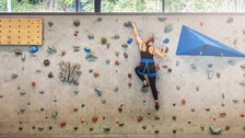 Foto: Eine Frau, die an einer Kletterwand klettert. Oberhalb der Kletterwand kann man durch Fenster ins Freie schauen. 