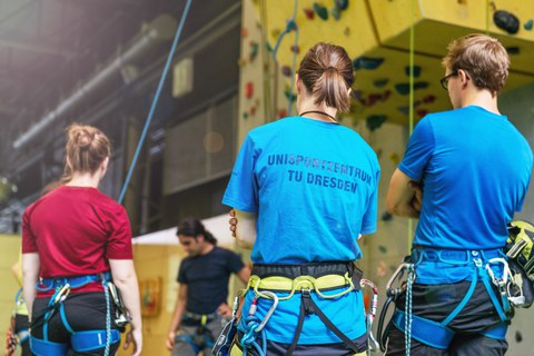 Foto von vier Personen in einer Kletterhalle. Drei von ihnen sind von hinten zu sehen. Zwei der Personen haben die Arme verschränkt. Alle tragen Klettergurte und bei einer Frau steht auf dem Rücken des Shirts Unisportzentrum TU Dresden. Im Hintergrund ist eine Kletterwand.