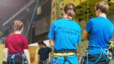 Foto von vier Personen in einer Kletterhalle. Drei von ihnen sind von hinten zu sehen. Zwei der Personen haben die Arme verschränkt. Alle tragen Klettergurte und bei einer Frau steht auf dem Rücken des Shirts Unisportzentrum TU Dresden. Im Hintergrund ist eine Kletterwand.