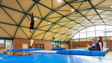 Foto: Ein Mann turnt in einer Sporthalle auf einem Trampolin. Zwei Frauen sitzen am Rand und schauen ihm dabei zu. 