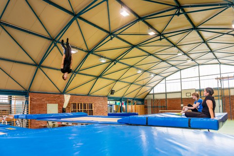 Foto: Ein Mann turnt in einer Sporthalle auf einem Trampolin. Zwei Frauen sitzen am Rand und schauen ihm dabei zu. 