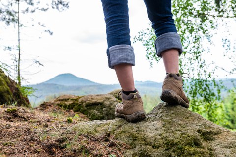 Man sieht die Beine einer Wanderin (knieabwärts), die auf einem Felsen steht, im Hintergrund ein anderer Felsen in der Sächsischen Schweiz.