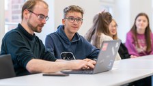 Teilnehmer schreibt am Laptop mit, ein weiterer Teilnehmer hört zu