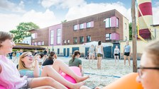 Es ist ein Foto von einem Beach-Volleyballplatz zu sehen. Im Vordergrund sitzen einige Mädchen und lachen. Im Hintergrund des Bildes stehen andere Mädchen im Spielfeld. 