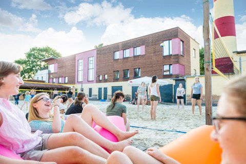 Es ist ein Foto von einem Beach-Volleyballplatz zu sehen. Im Vordergrund sitzen einige Mädchen und lachen. Im Hintergrund des Bildes stehen andere Mädchen im Spielfeld. 