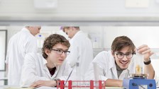 Es ist ein Foto zu sehen. Vier Personen befinden sich in einem Labor. Sie tragen weiße Kittel. Zwei männliche Studieninteressierte sitzen im Vordergrund an einem Tisch und experimentieren. Im Hintergrund stehen ebenfalls zwei Jugendliche.