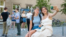 Zwei Schülerinnen sitzen auf der Bank vor dem Gerhart-Potthoff-Bau, lächeln und machen ein Selfie. Im Hintergrund stehen weitere Schülerinnen.
