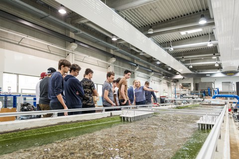 Zu sehen ist ein Foto. Eine Jungengruppe steht in einer großen Halle hinter dem Modell eines Flusses. Die Blicke sind auf das Modell gerichtet. Eine Frau zeigt auf dieses Modell und spricht. 