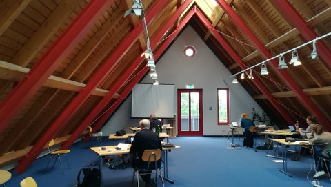 Foto Seminarraum mit spitzer Decke. Tische, an denen schreibende Personen sitzen (Rückenansicht).