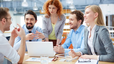 A group of five people in an office are gathered around a table and talking. On the table there is an open laptop as well as papers, pens and glasses.
