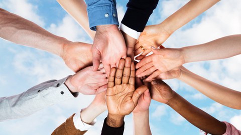 Photo of many outstretched hands of different skin colors lying crossed over each other, in the background is the sky