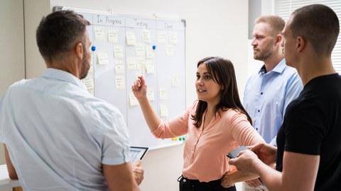 Four people are gathered at a whiteboard in an office. The board shows a project planning system.