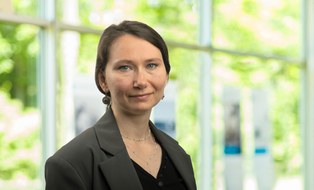Porträtfoto. hellgrüner Hintergrund. Frau Kuzina trägt langes, zurückgebundenes Haar. Ihre rechte Schulter ist nach vorn gedreht und sie trägt einen hellgrauen Blazer.