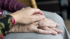 Auf dem Foto eine alte Hand, die aufs Knie geleget ist. Darunter die andere Hand. Fürsorglich liegt, von der Seite kommend, eine jüngere Hand darüber.