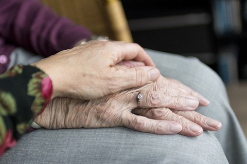 Auf dem Foto eine alte Hand, die aufs Knie geleget ist. Darunter die andere Hand. Fürsorglich liegt, von der Seite kommend, eine jüngere Hand darüber.