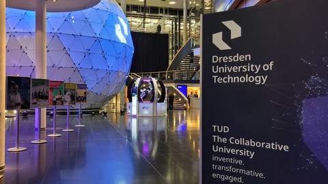 Modernes Foyer mit einer großen, blau leuchtenden geodätischen Kuppel im Hintergrund. Im Vordergrund stehen eine Informationstafel einer Universität sowie eine Reihe von Ausstellungsplakaten auf Ständern.