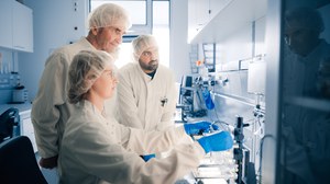 Drei Forschende in Laborkitteln und Kopfbedeckungen arbeiten an wissenschaftlichen Geräten in einem sauberen, modernen Labor.
