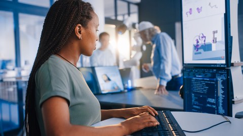 Eine fokussierte, junge Frau mit Zöpfen und einem grünen T-Shirt tippt auf einer Tastatur. Sie blickt auf zwei Monitore, auf denen Computercode und ein 3D-Modell einer Roboterhand zu sehen sind. Im Hintergrund interagieren zwei weitere Personen.