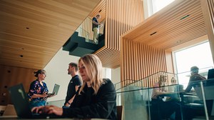  Mehrere Personen arbeiten in einem modernen, mehrstöckigen Atrium mit Holzlamellen und Glasgeländern. Eine junge Frau im Vordergrund tippt auf einem Laptop, während andere sich unterhalten, auf einer oberen Etage stehen oder an Tischen sitzen.